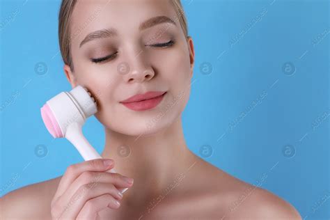 Young woman washing face with cleansing brush on light blue background. Cosmetic product: Stock ...