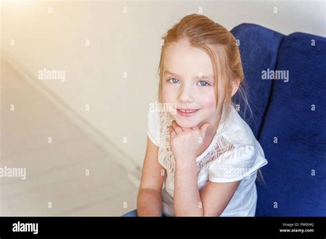 Sweet little girl in jeans and white T-shirt at home sitting on modern cozy blue chair relaxing ...