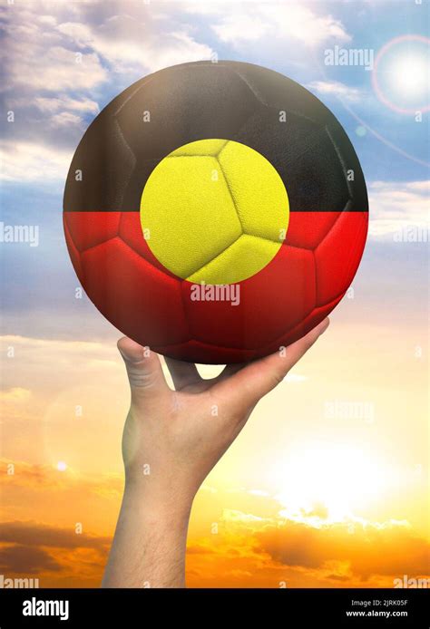 Soccer ball in hand with a depiction of the flag of Australian Aboriginal against a colorful sky ...