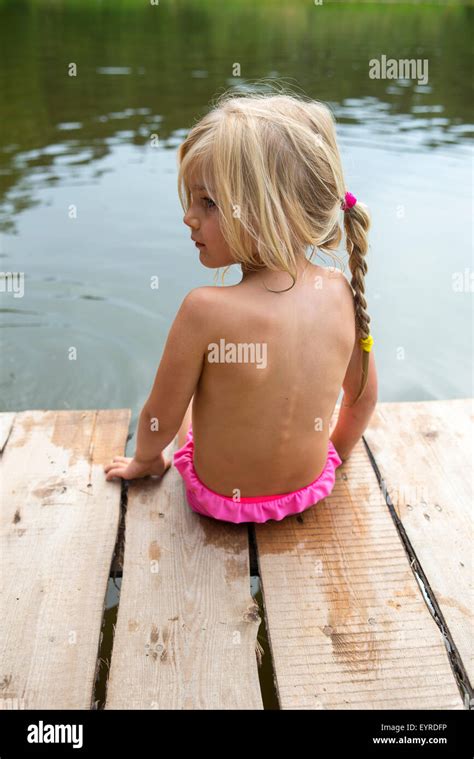 Rear view of child blond girl sitting on pier looking at view of pond with her feet in the water ...