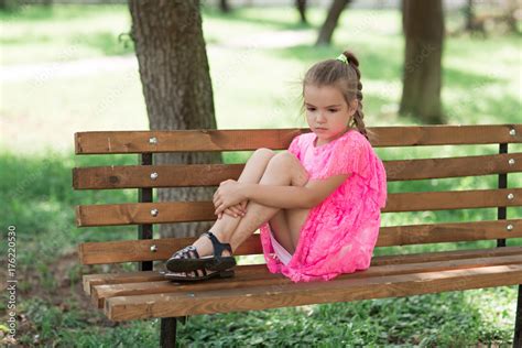 Little caucasian girl sitting in park on bench. A child in a beautiful pink dress in the open ...