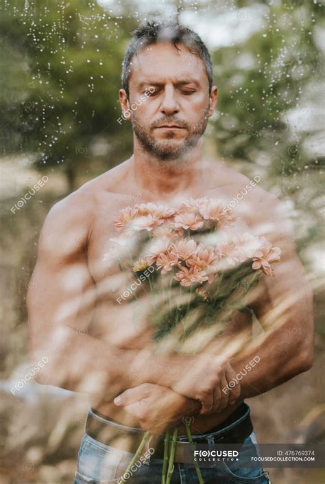 Shirtless handsome man with eyes closed holding bouquet in forest seen through glass — Mid Adult ...