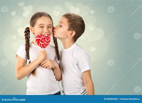 Мальчик целует маленькую девочку с леденцом на палочке конфеты красным ...