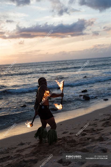 Male fire dancer performing with fire levi sticks at beach in twilight