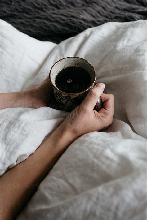 "Two Hands Holding A Mug Of Coffee On A White Bedspread" by Stocksy