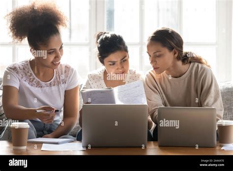 Diverse girls studying together at home, reading notes, using laptops ...