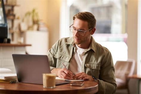 Imagen de un joven enfocado con gafas sentado en un café tomando notas ...