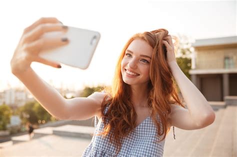 Улыбающаяся рыжая женщина с длинными волосами, делающая селфи ...