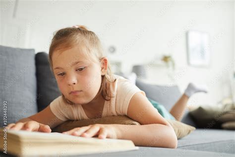Little girl with down syndrome lying on sofa and reading a book in the