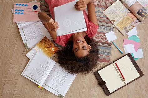 Top view of attractive young curly female with dark skin lying on carpet while reading notes ...