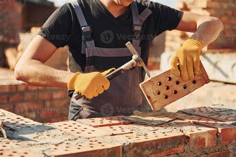 Holding brick and using hammer. Construction worker in uniform and