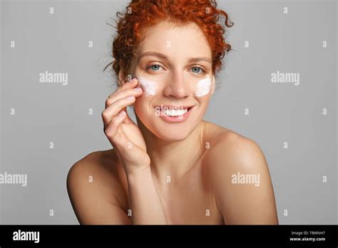 Beautiful redhead woman applying facial cream against grey background