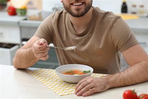 Молодой человек ест вкусный овощной суп за столом на кухне, крупным ...