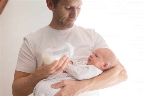 A father holding and feeding his newborn son with milk bottle at home ...